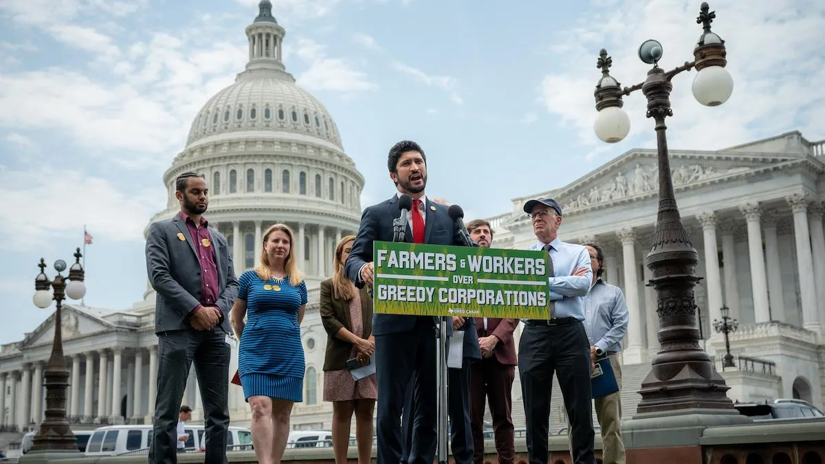 Texas Rep. Greg Casar speaks at a podium with a group of people behind him. The podium has a sign reading "Farmers & workers over greedy corporations."