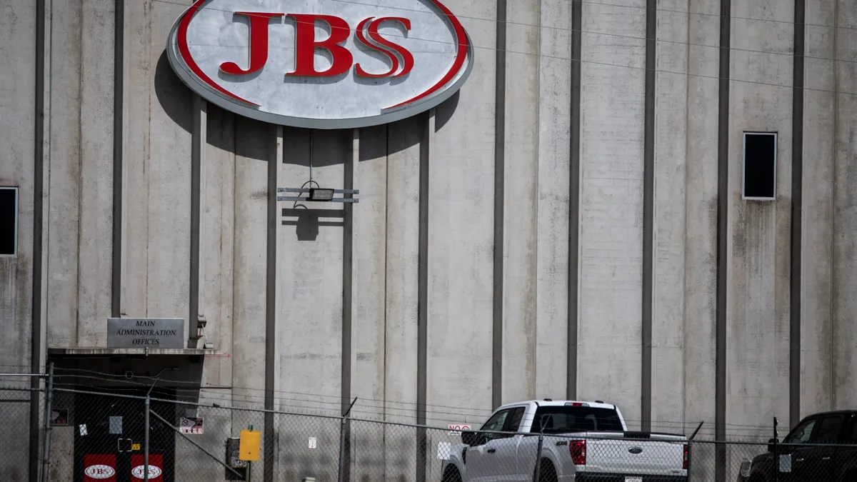 The outside of a factory is seen. A sign with "JBS" in red letters is plastered on the front.