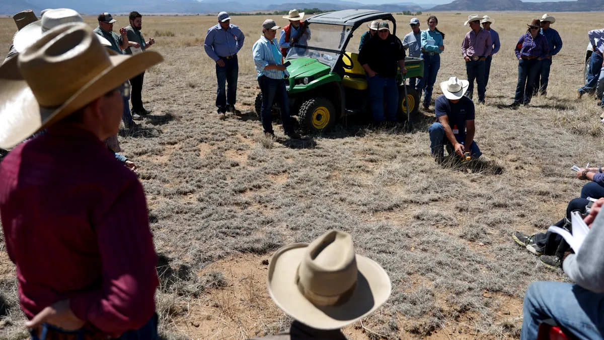 A group of people in cowboy hats loosely stand in a circle watching as a man takes soil temperature