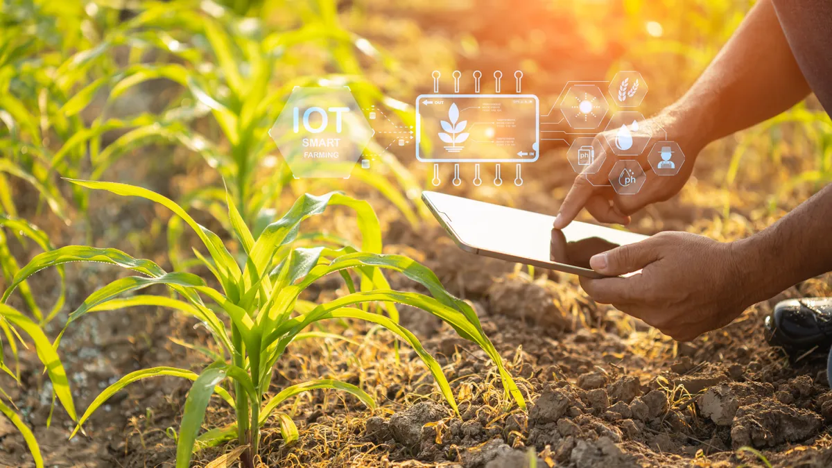 A hand holding a tablet near a farm field is seen.