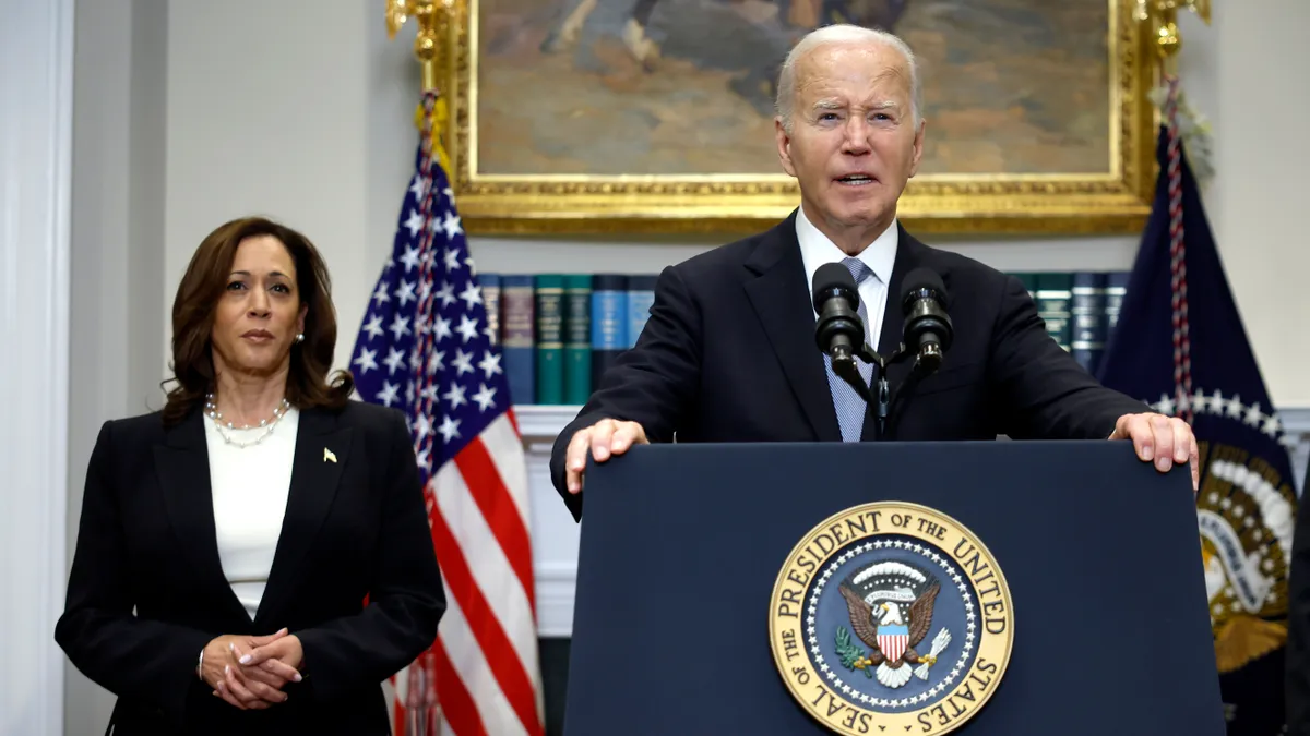 A person in a suit stands at a podium with a microphone. A woman stands behind him with hands clasped in front of her. Behind them are flags.