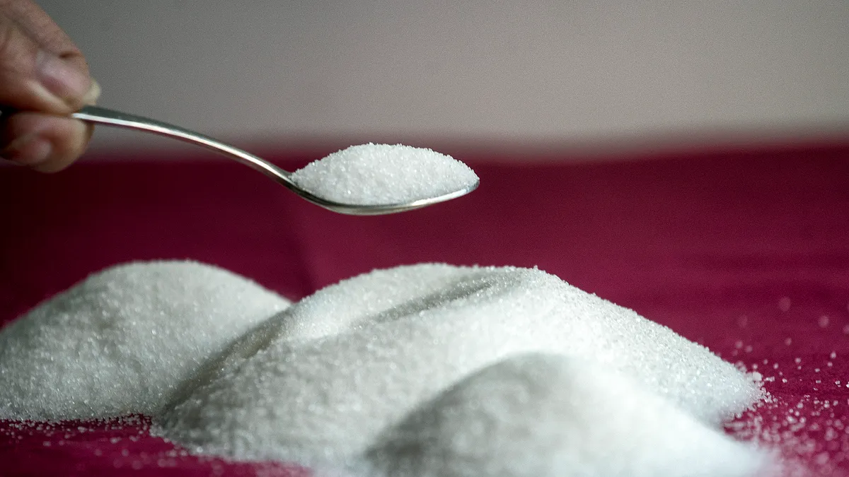 Mounds of sugar are on a red surface. A hand holds a teaspoon full of sugar over one of them.