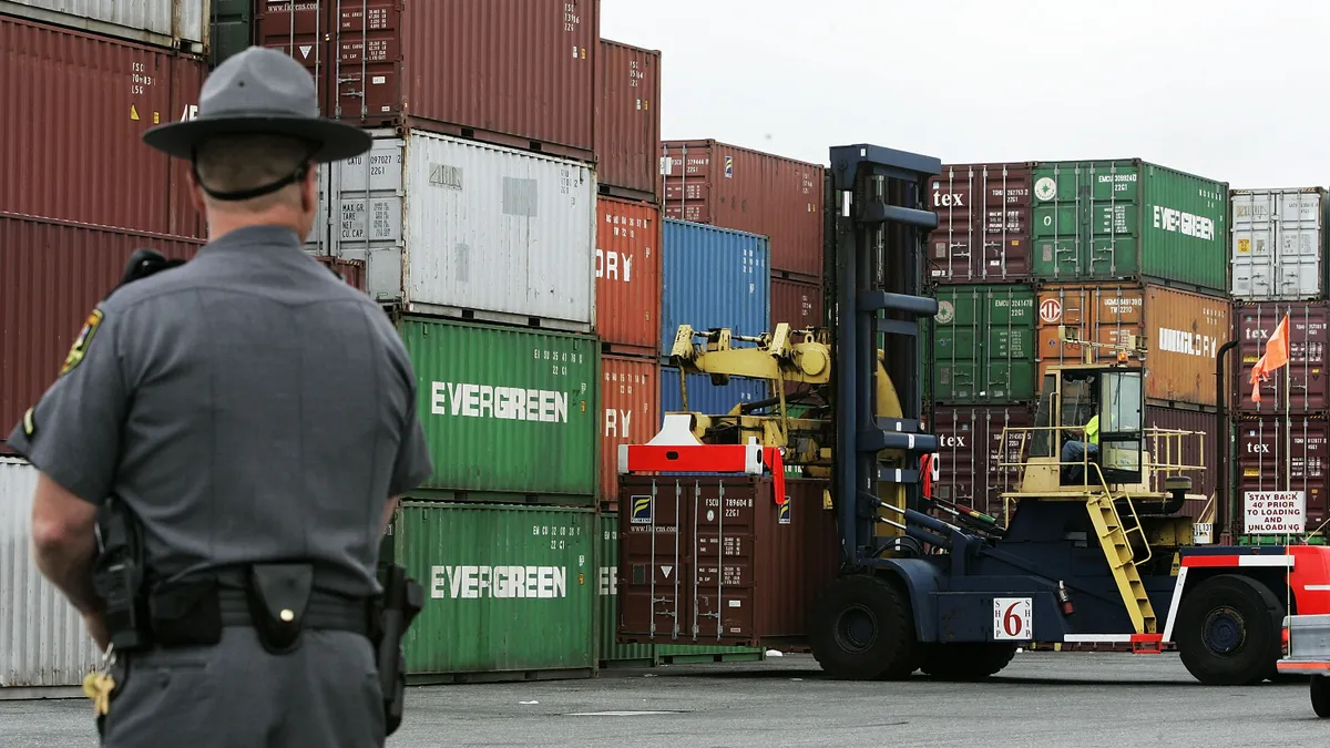 An officer is seen from behind as he looks at a stack of ocean shipping containers.