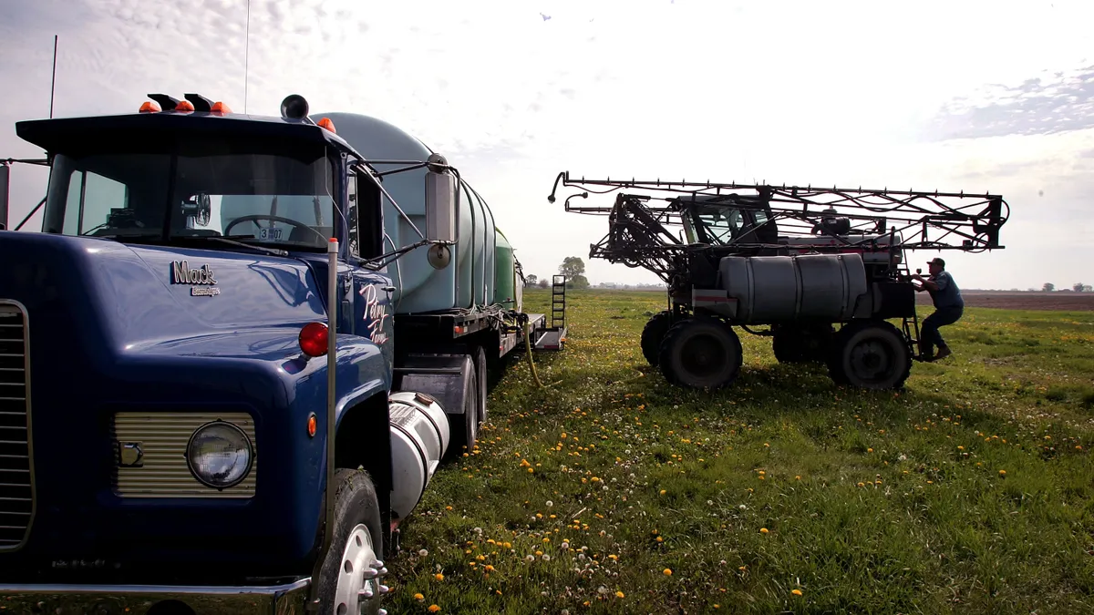 A farmer stands next to an herbicide sprayer. Next to him is a truck.