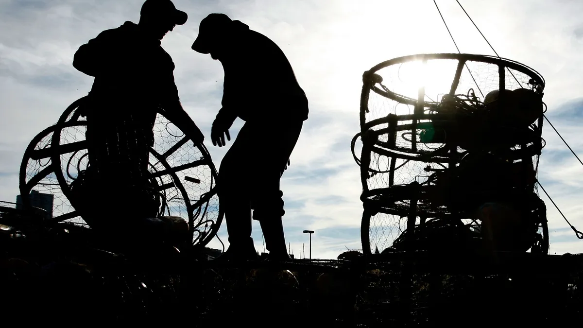 The silhouettes of two fishermen on a boat are seen as they load circular crab traps onto a boat