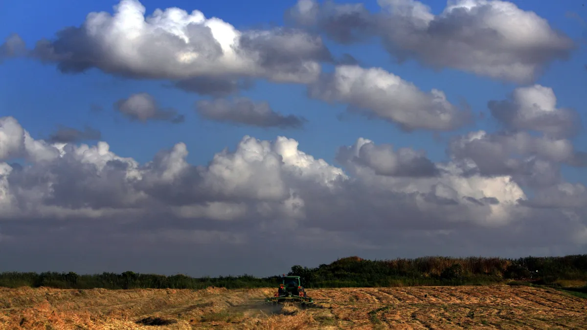 A tractor turns hay in a field under a partly cloudy sky.