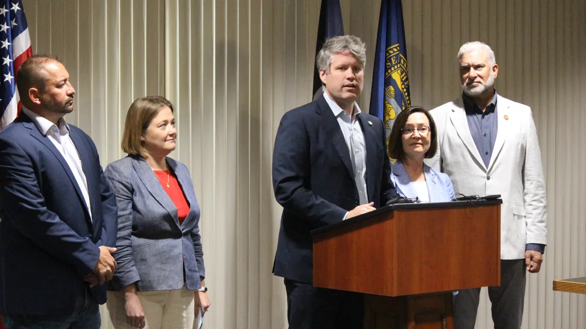 A man speaks at a podium with a group of people surrounding him