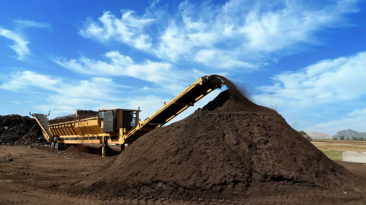 Earthy material is deposited from a wheeled machine's large conveyor belt onto an outdoor pile.